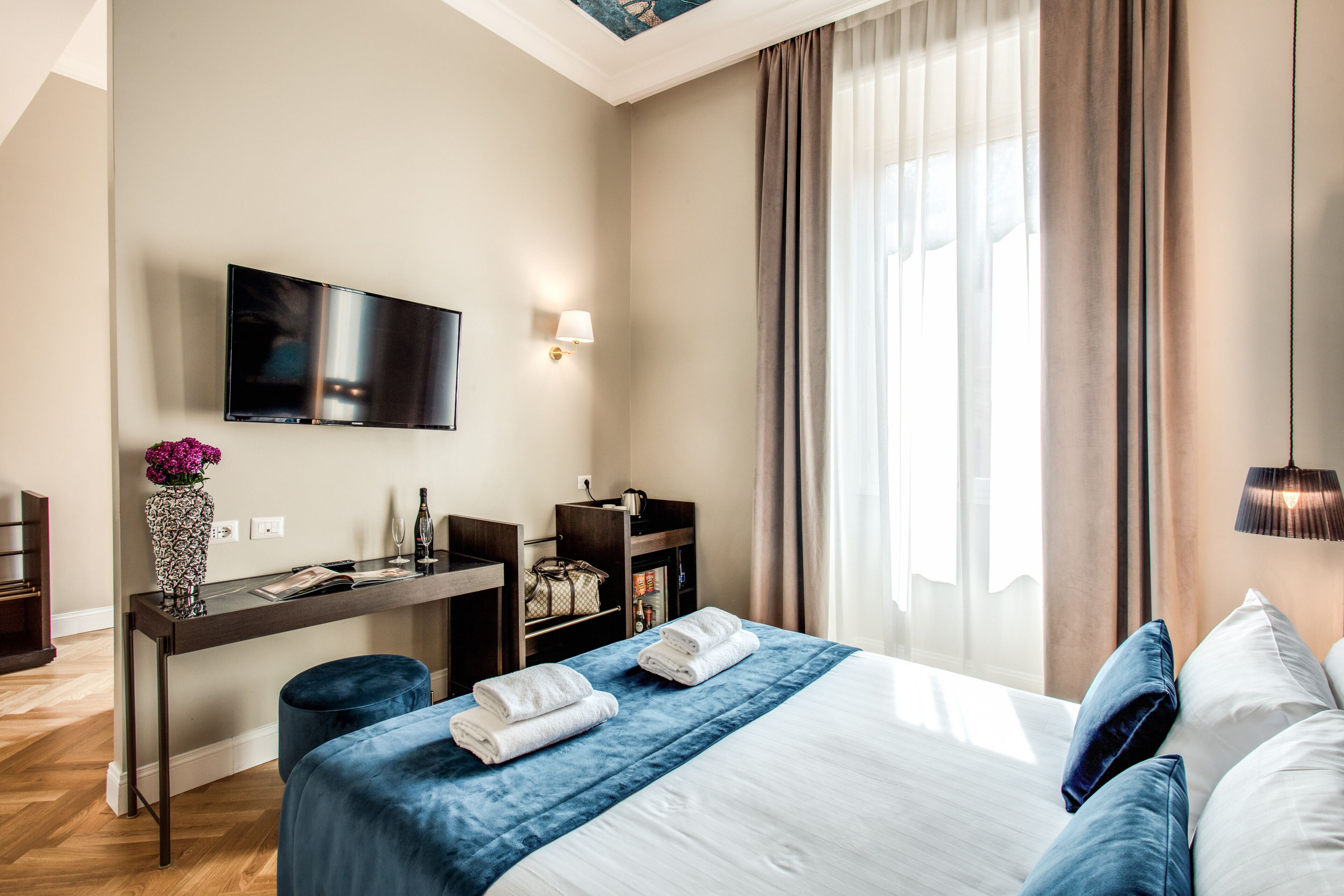 A hotel room with a bed, a desk with a flat-screen TV, a vase with flowers, and a large window with curtains.