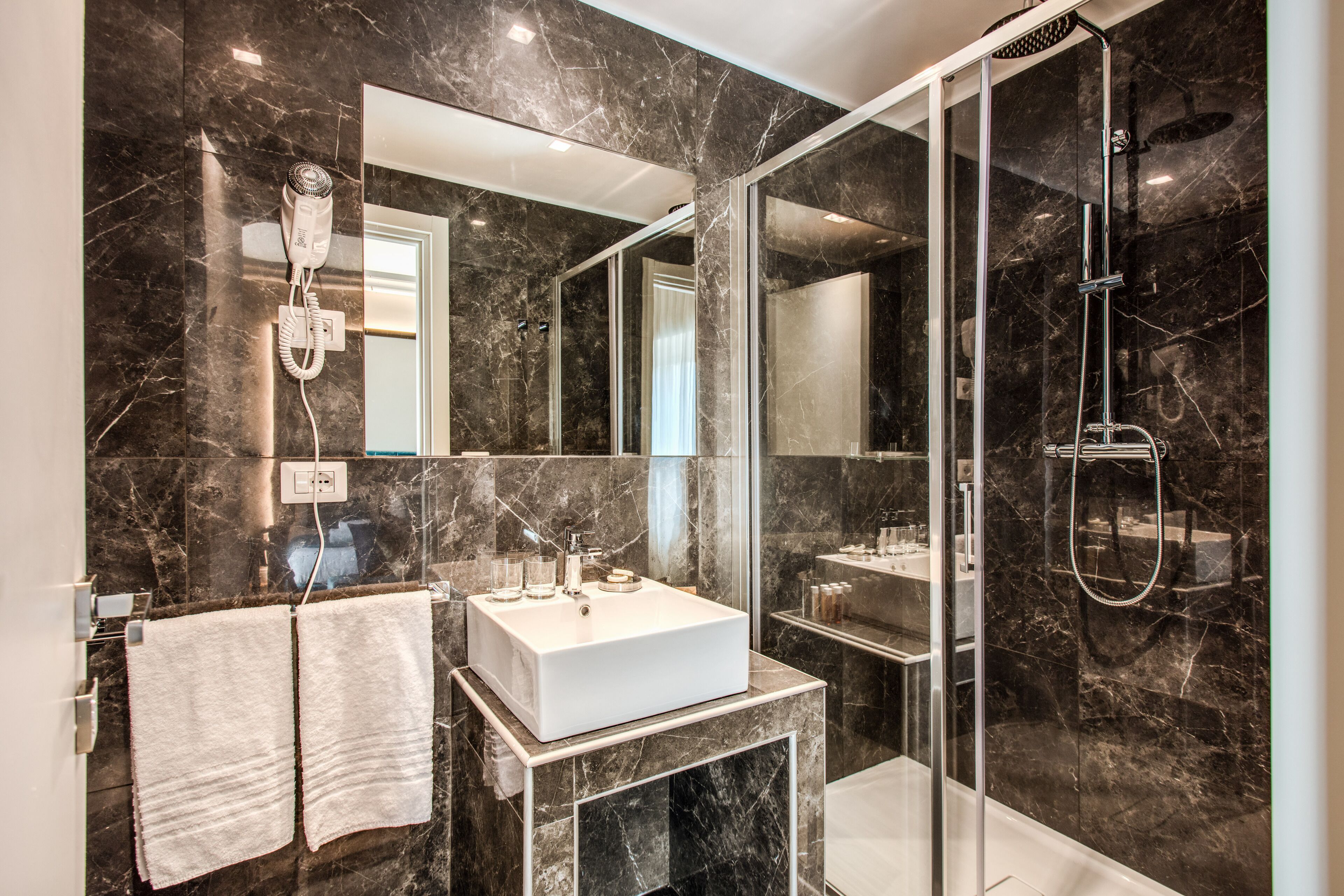 A modern bathroom with a large mirror, a white sink, a glass shower enclosure, and a hairdryer mounted on the wall.