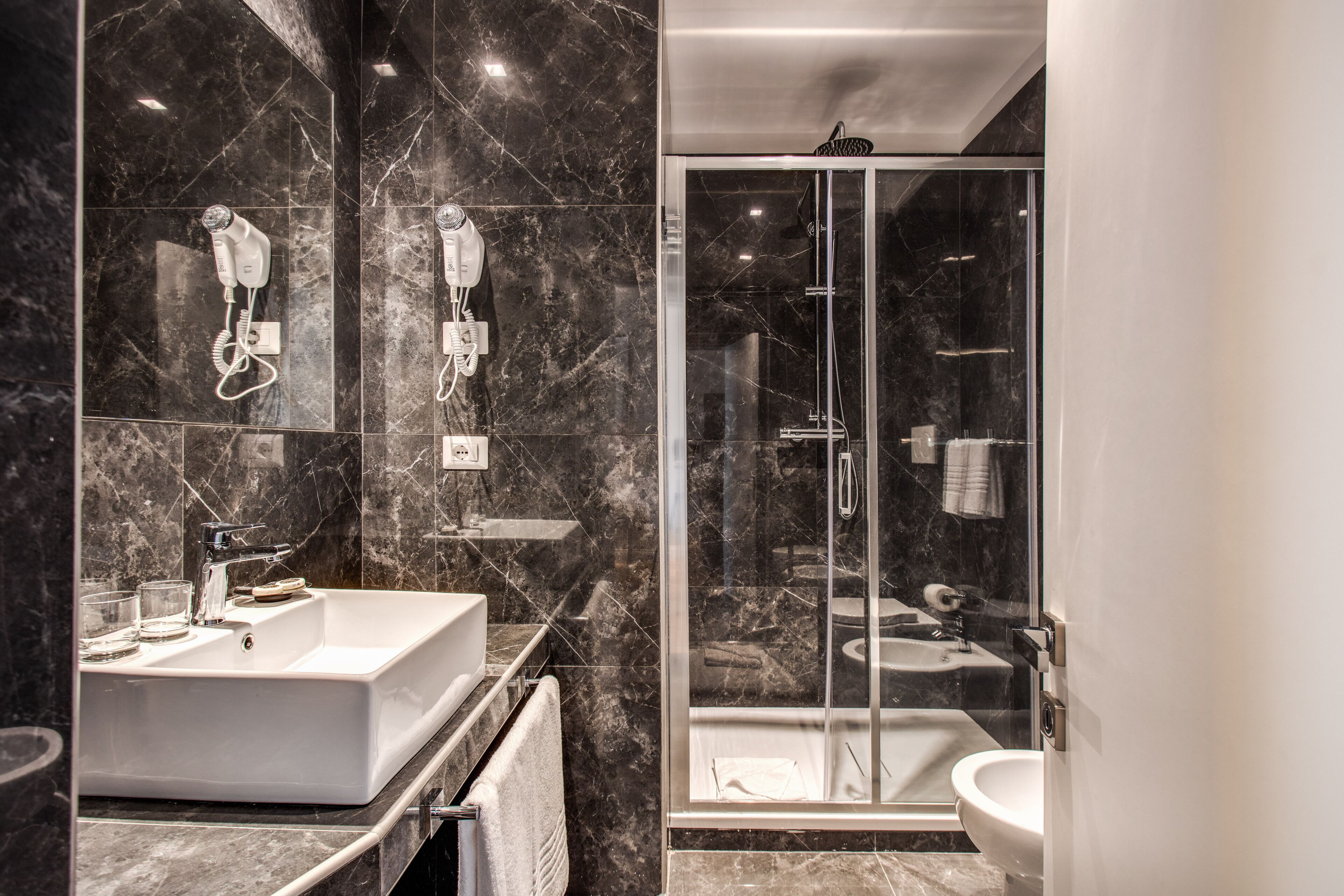 A modern bathroom with a large white sink, a glass-enclosed shower, and two hair dryers mounted on the wall.