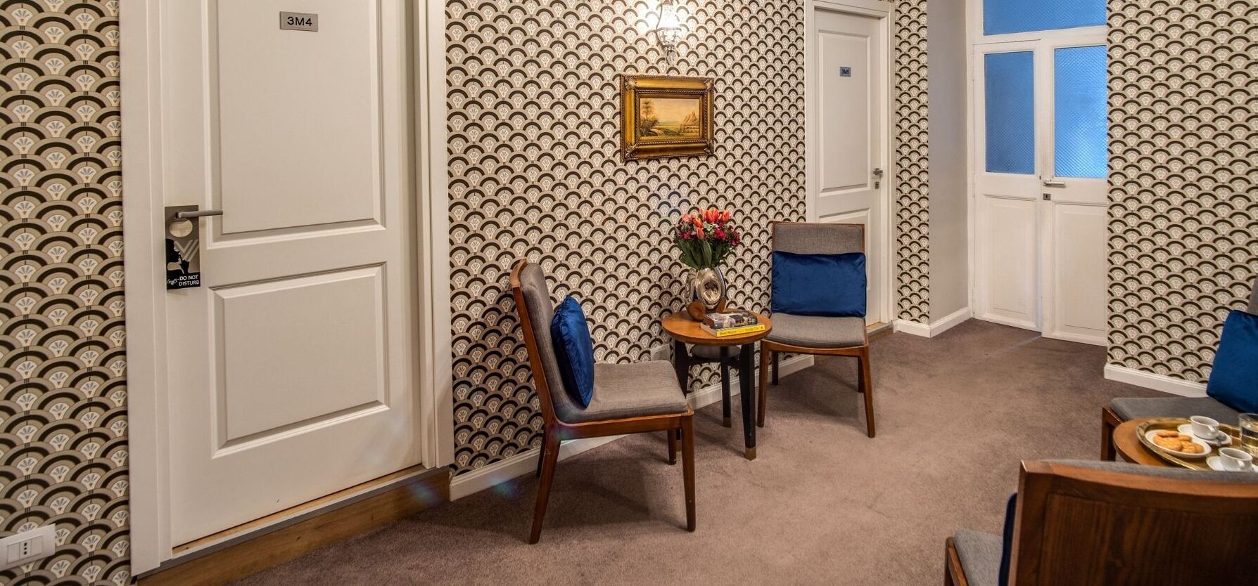 A hallway with patterned wallpaper, a framed picture on the wall, and a table with a vase of flowers.