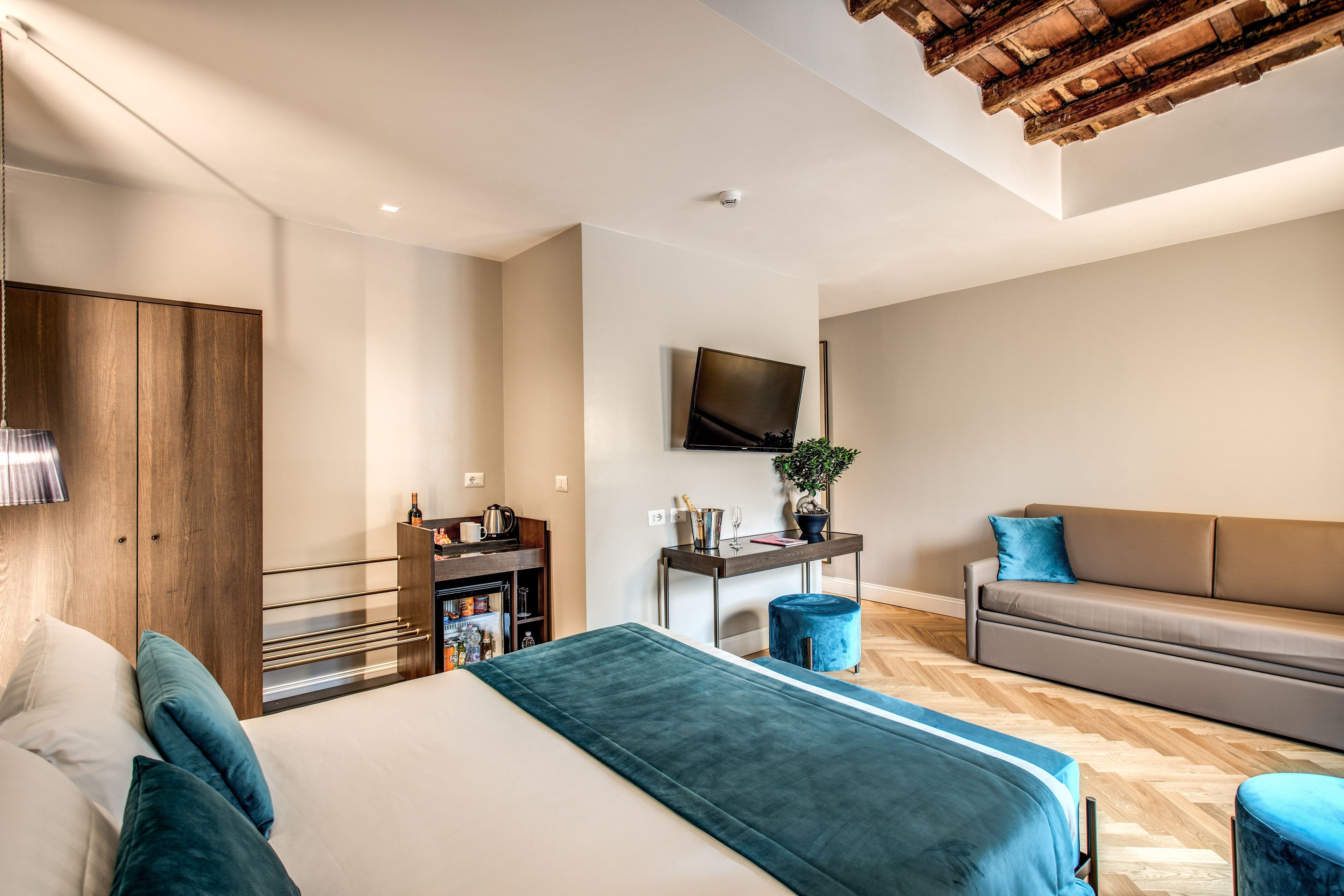 A modern hotel room with a large bed, a sofa, a TV mounted on the wall, and a wooden ceiling.