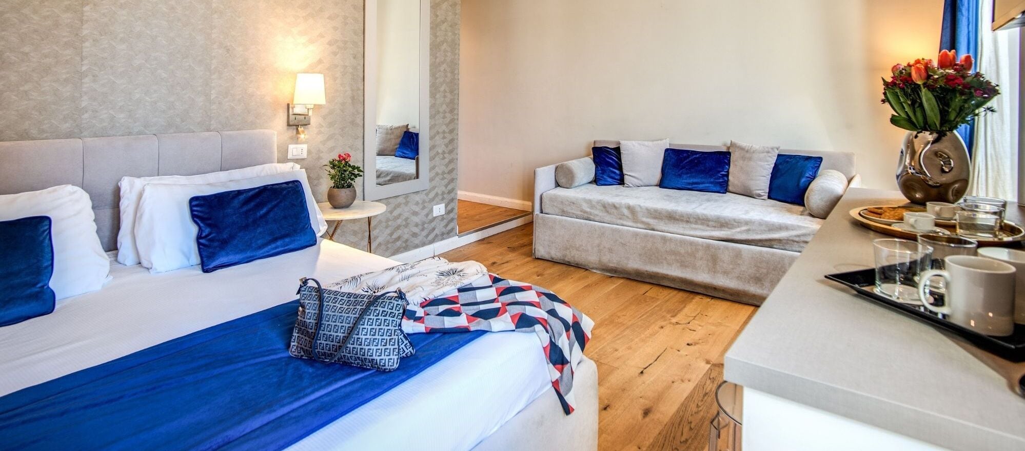 Une chambre d’hôtel avec deux lits, un canapé, une petite table sur laquelle se trouve un plateau avec des tasses, et un vase rempli de fleurs.