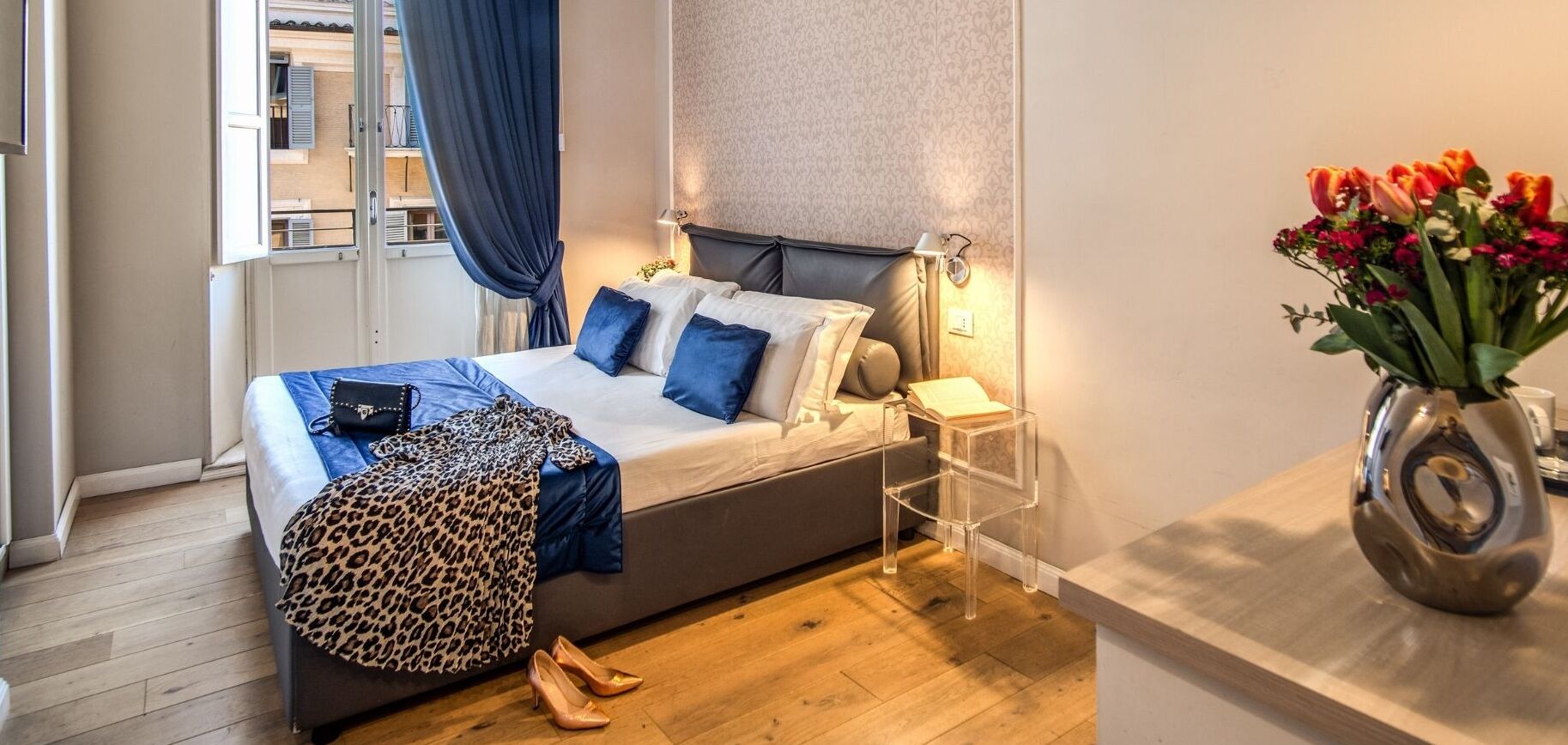 A bedroom with a bed, bedside table, lamp, and a vase with flowers.