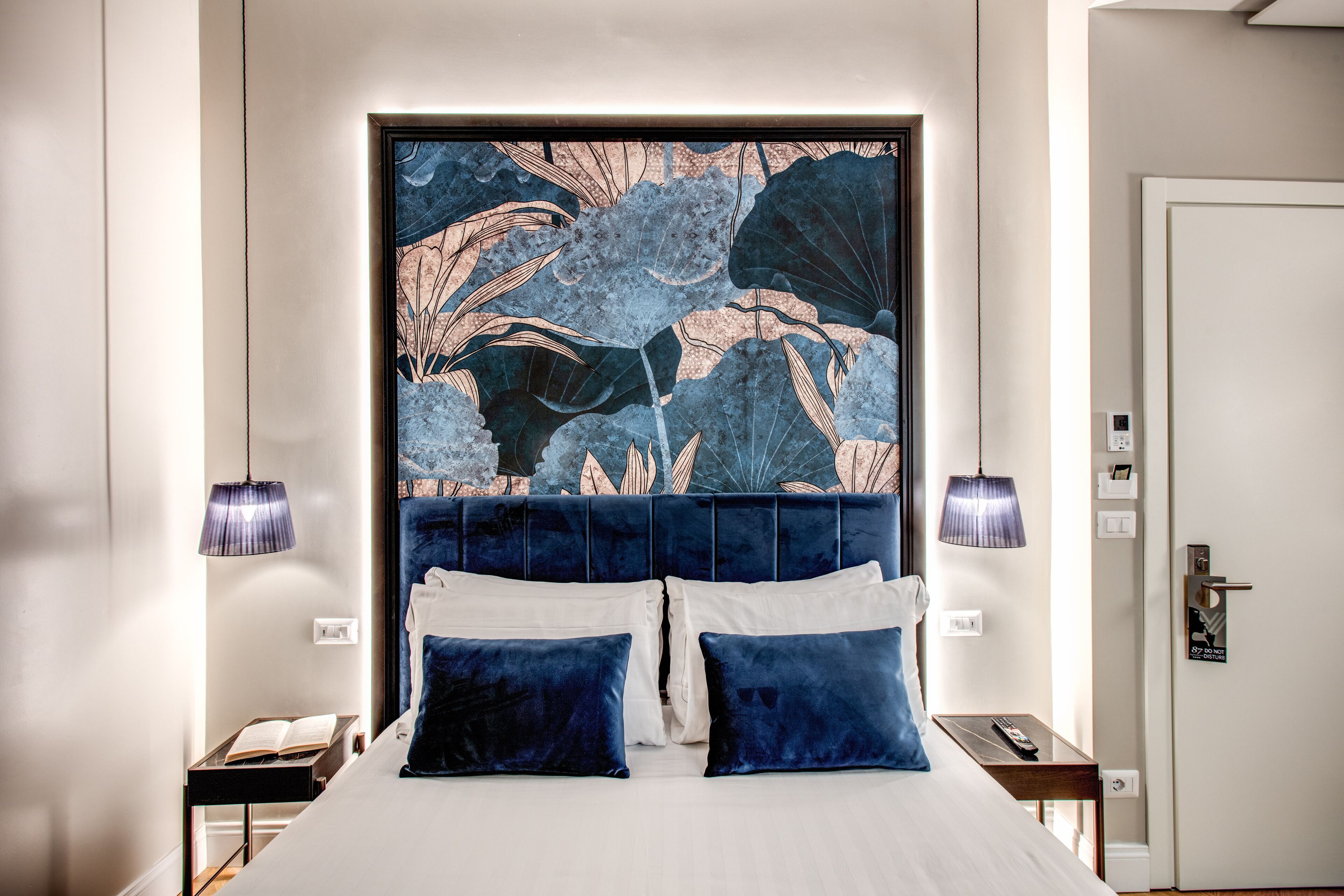 A neatly made bed with a headboard featuring a large, detailed artwork. There are two bedside tables with lamps, and a door with a handle and lock.