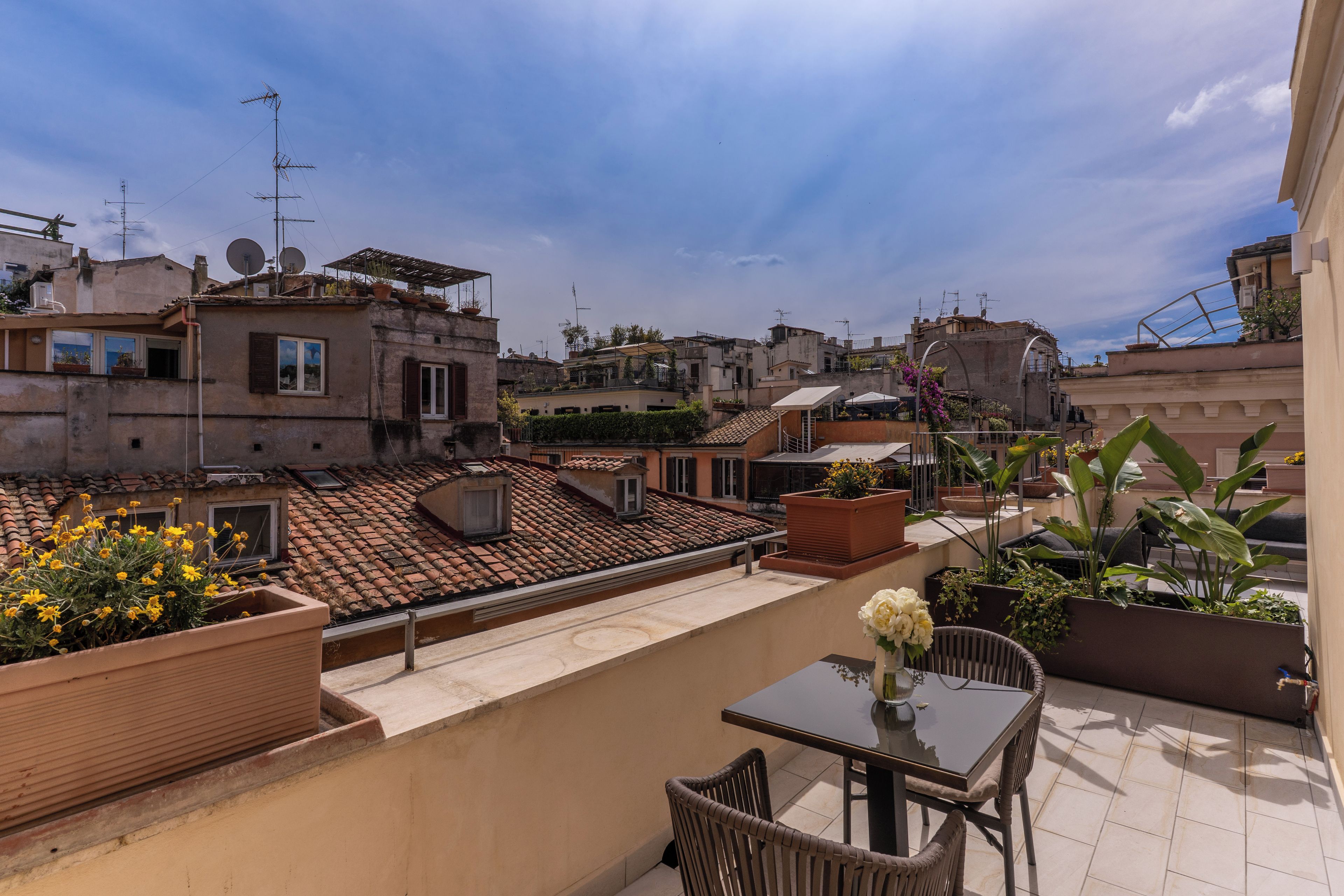 A rooftop terrace with potted plants, a table, and chairs.