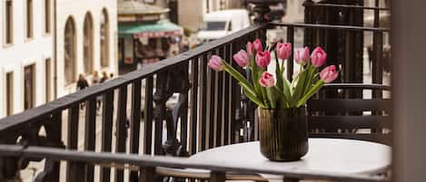 En balkong med ett bord och stolar, en vas med rosa blommor och utsikt över en stadsgata med byggnader.