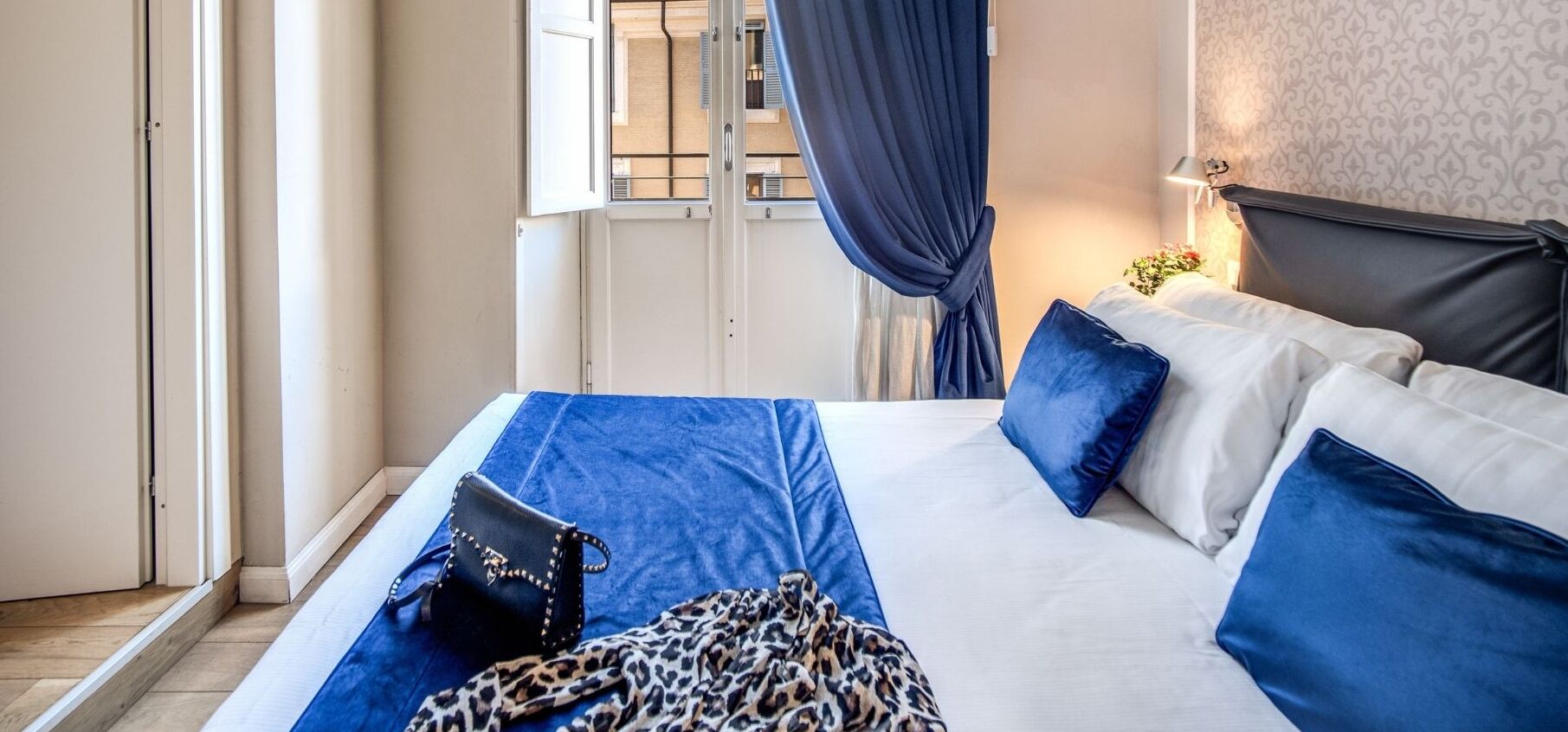 A hotel room with a bed, a nightstand, a lamp, a window with curtains, and a view of the outside.