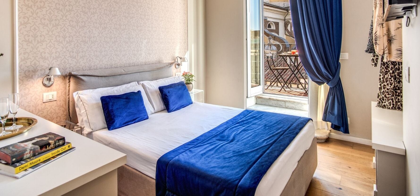 A neatly made bed with blue bedding, a bedside table with a book, and a window with a view of a balcony.