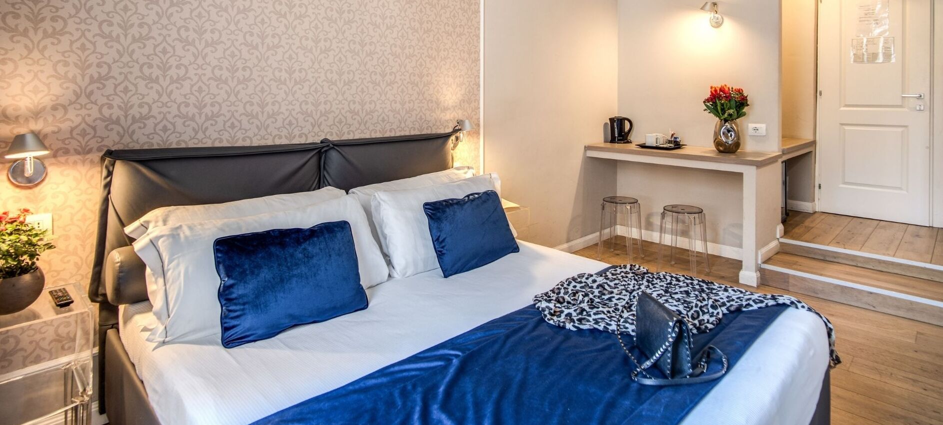 Un lit bien fait, avec une tête de lit noire, du linge de lit blanc et des coussins bleus. On y trouve une petite table sur laquelle sont posés un vase de fleurs et une bouilloire.