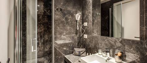 A modern bathroom with a glass-enclosed shower, a marble-tiled wall, a large mirror, and a vanity with a sink and toiletries.