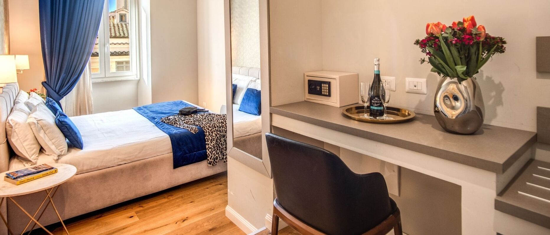 A hotel room with a bed, a desk with a vase of flowers, a remote control, and a bottle of wine.