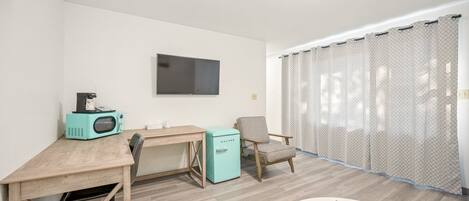 A compact room with a wooden desk, a teal-colored refrigerator, a chair, a TV mounted on the wall, and a window with sheer curtains.
