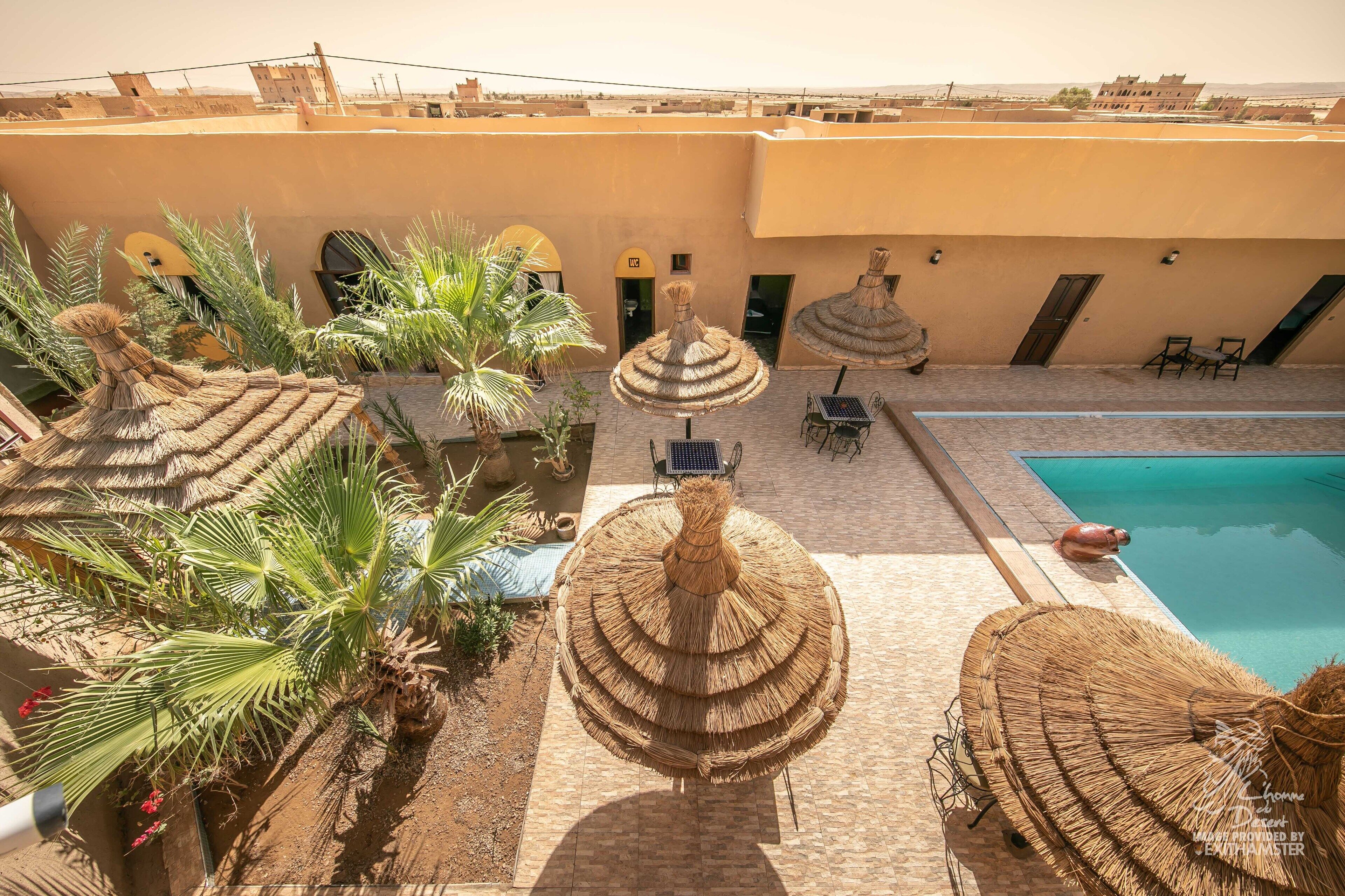 A rooftop pool area with thatched umbrellas, palm trees, and a view of the surrounding buildings.