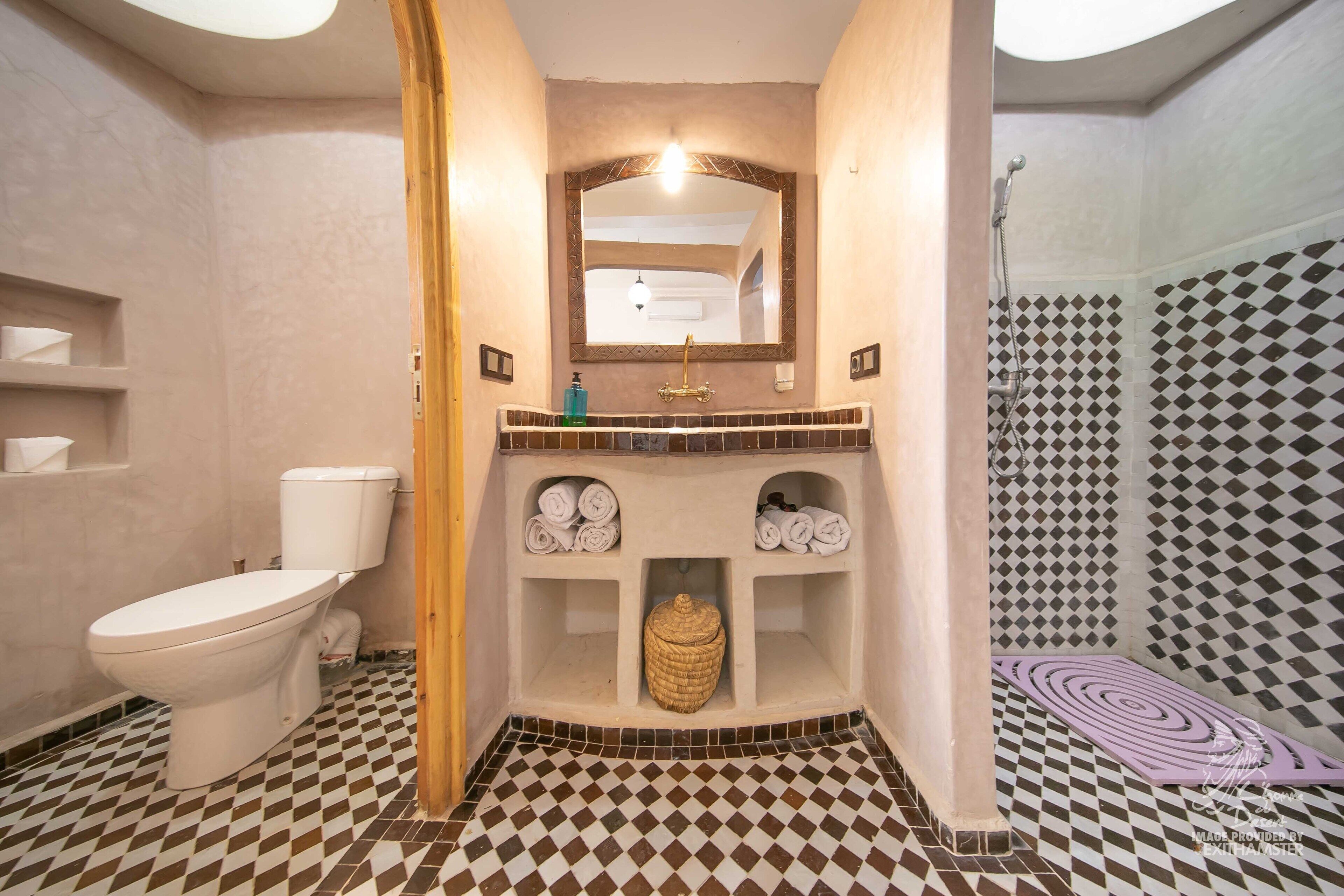 Casa de banho com sanita, lavatório e chuveiro. O chão tem um padrão quadriculado e há prateleiras com toalhas.