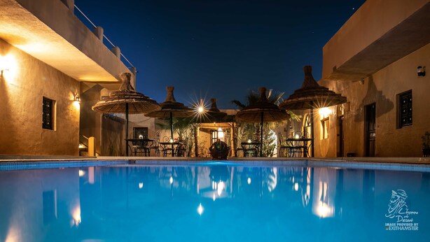 A nighttime pool area with thatched umbrellas, tables, and chairs, surrounded by buildings with warm lighting. - L'Homme du Désert (Taouz)