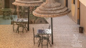 A courtyard with thatched umbrellas, tables, and chairs, surrounded by palm trees and a view of sand dunes. - L'Homme du Désert (Taouz)