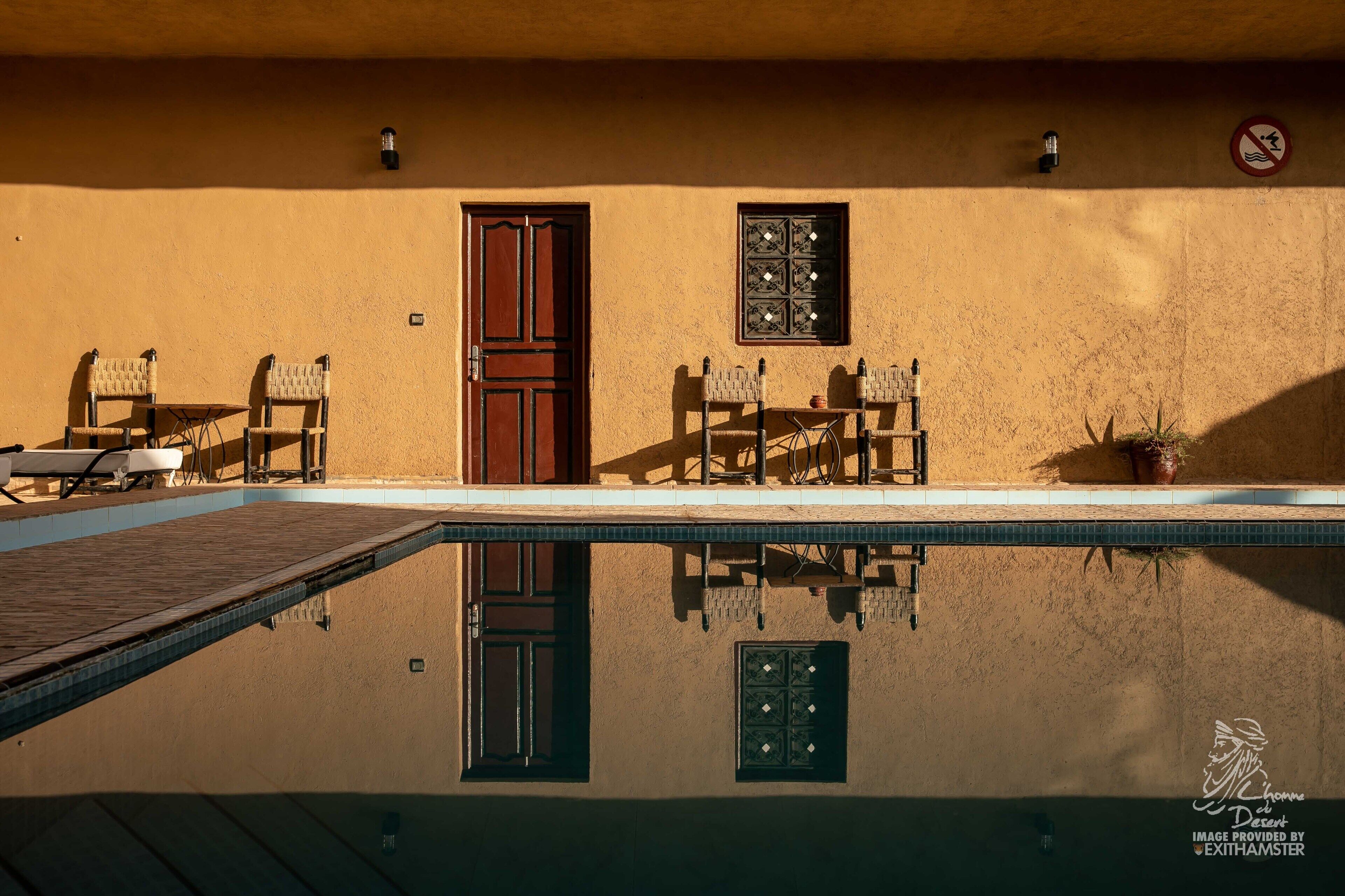 Zona de piscina con el reflejo de una puerta y una ventana, dos sillas y una planta en maceta.