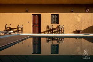 Zona de piscina con el reflejo de una puerta y una ventana, dos sillas y una planta en maceta.