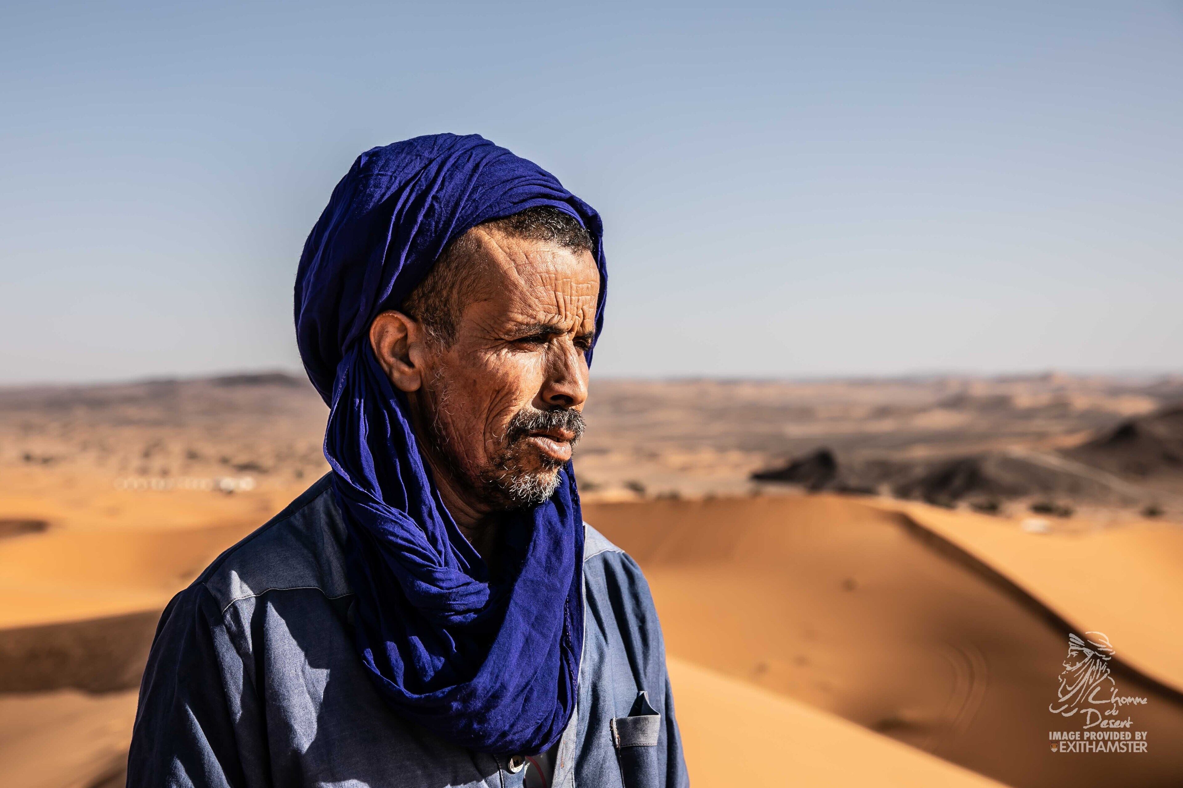 A person in a blue headscarf standing in a desert landscape with sand dunes in the background.