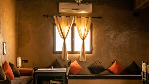 A cozy room with a bench, a table, and a chair. There is a window with curtains and an air conditioning unit above it. - L'Homme du Désert (Taouz)