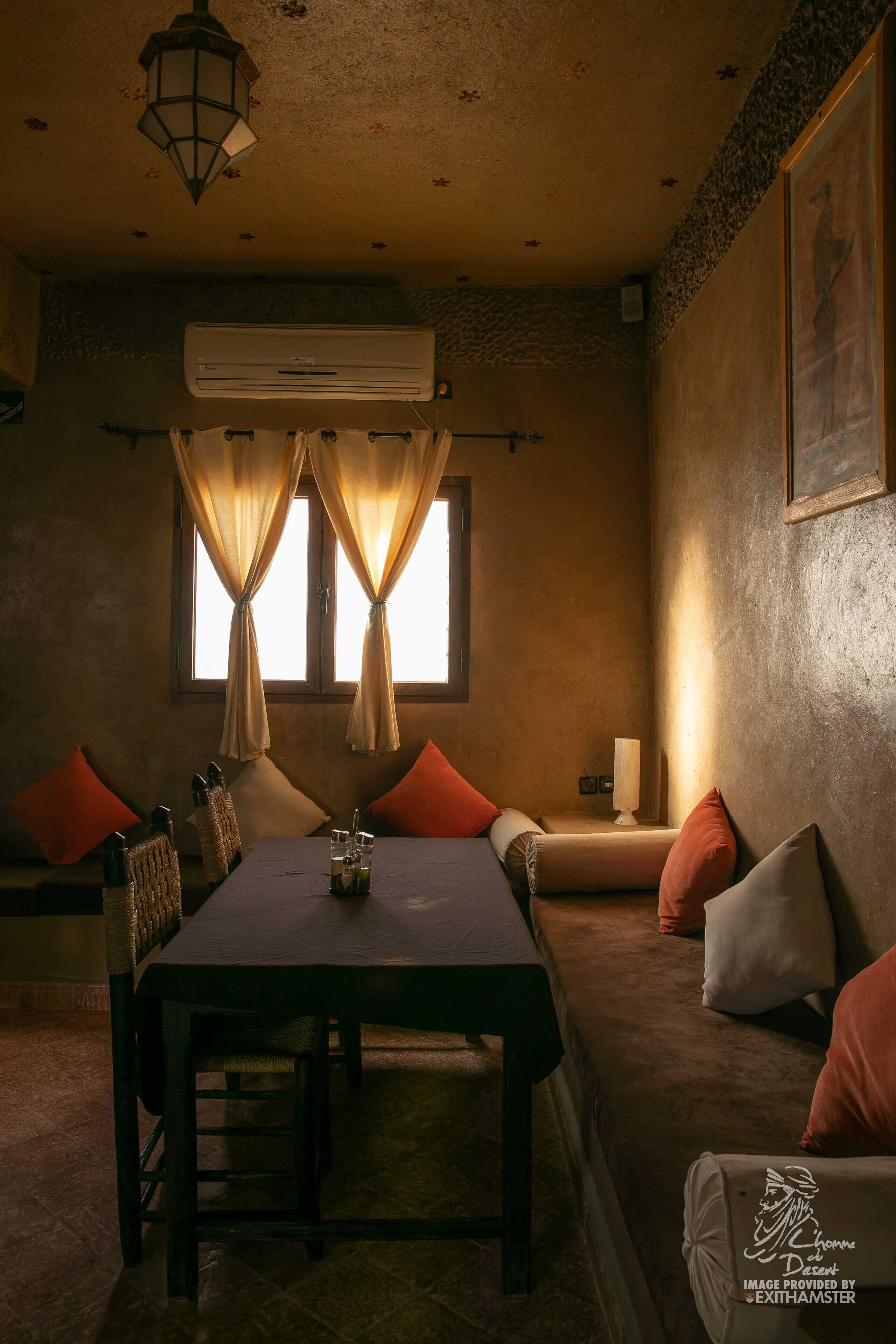 A dining area with a table, chairs, and a sofa. There is an air conditioning unit on the wall, a window with curtains, and a wall-mounted light.