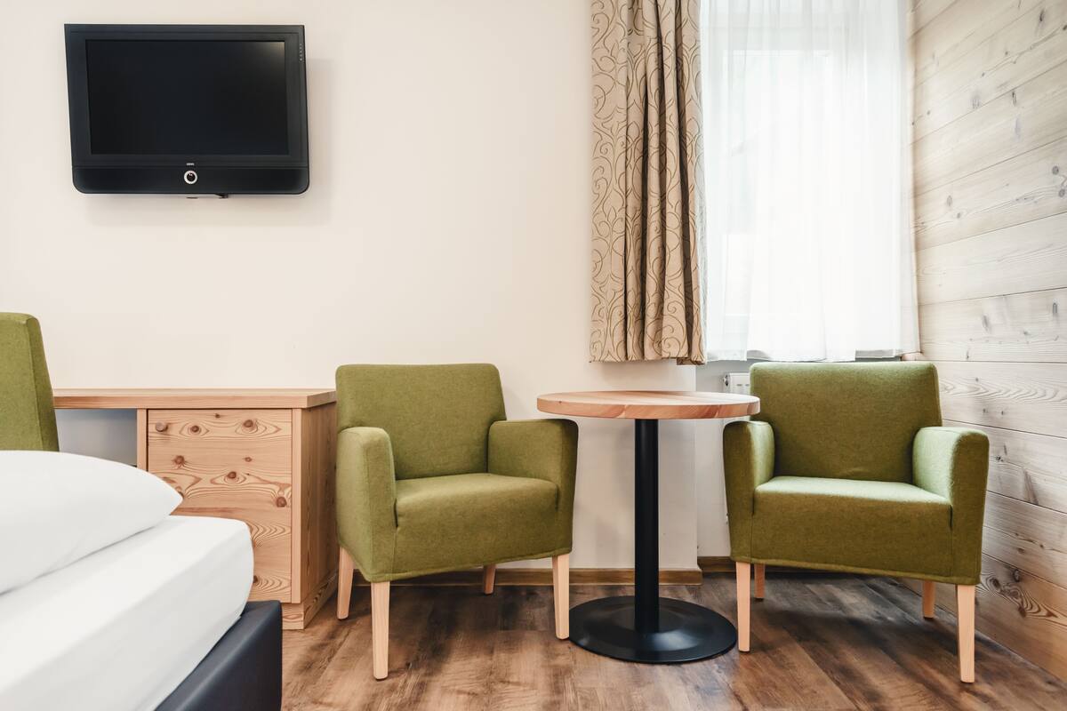 Ein Hotelzimmer mit einem Bett, einem Schreibtisch, zwei grünen Sesseln, einem runden Tisch, einem an der Wand montierten Fernseher und einem Fenster mit Vorhängen.