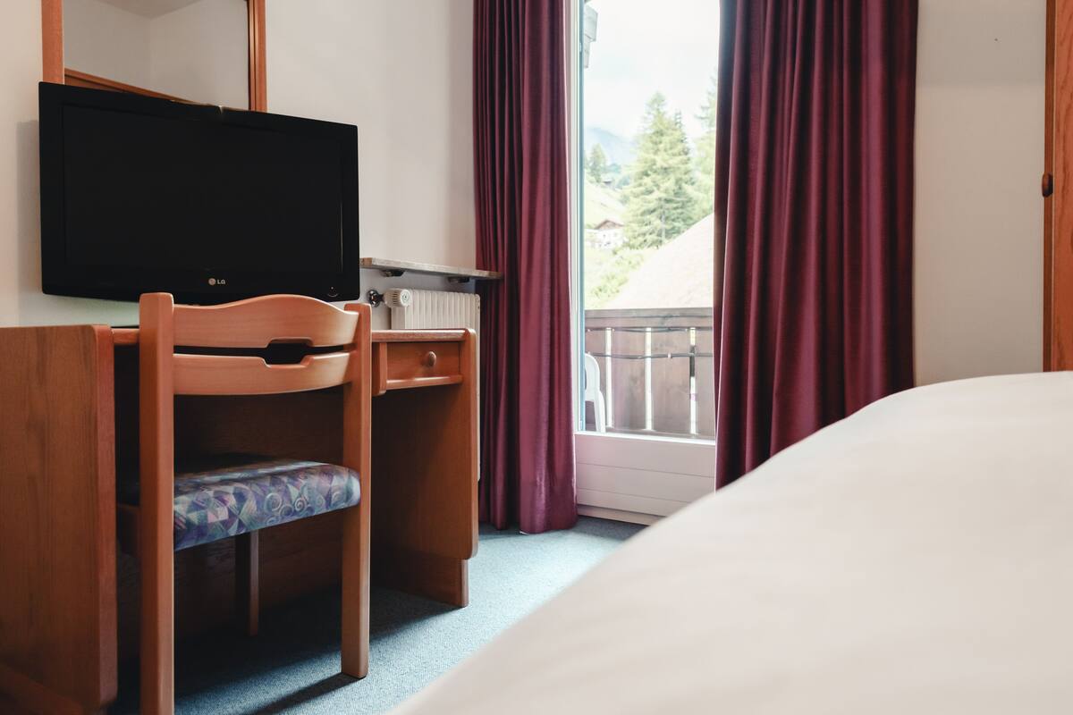 Ein Hotelzimmer mit einem Bett, einem Holztisch mit Stuhl, einem Fernseher und einem Fenster mit Vorhängen.