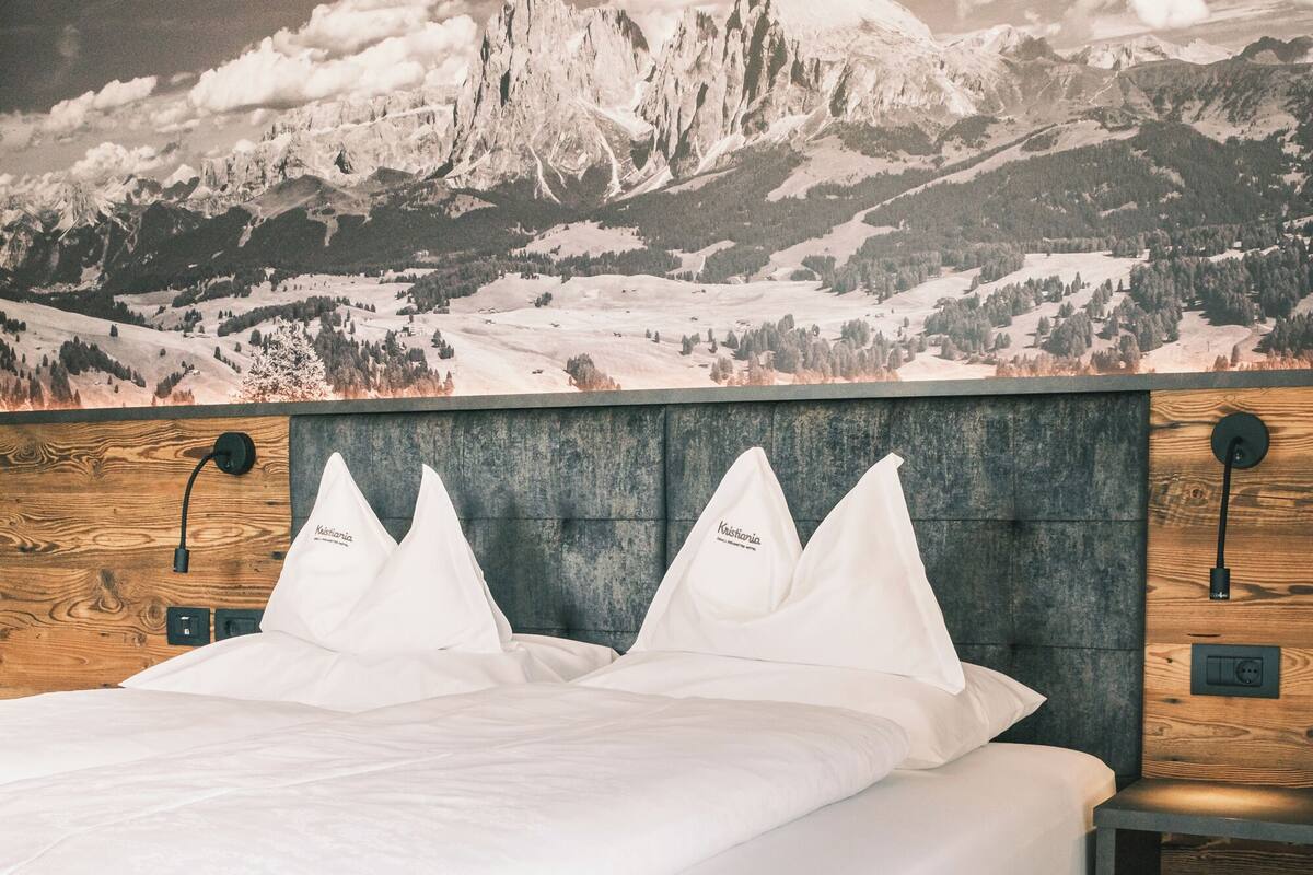 Ein ordentlich bezogenes Bett mit weißer Bettwäsche und Kissen, ein Holz-Kopfteil und ein Nachttisch mit Lampe.