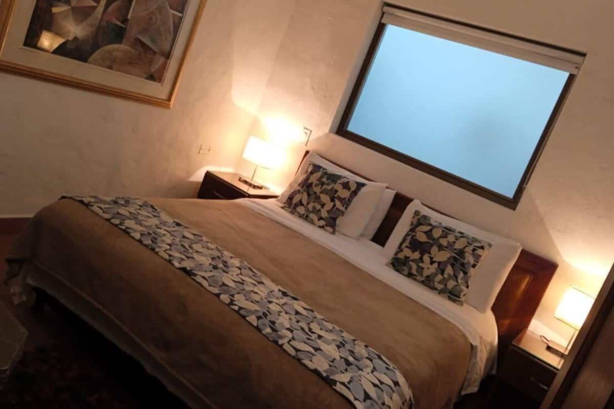 Une chambre d’hôtel avec un grand lit, une table de chevet, une lampe de chevet, un tableau encadré au mur et une fenêtre donnant sur un paysage.
