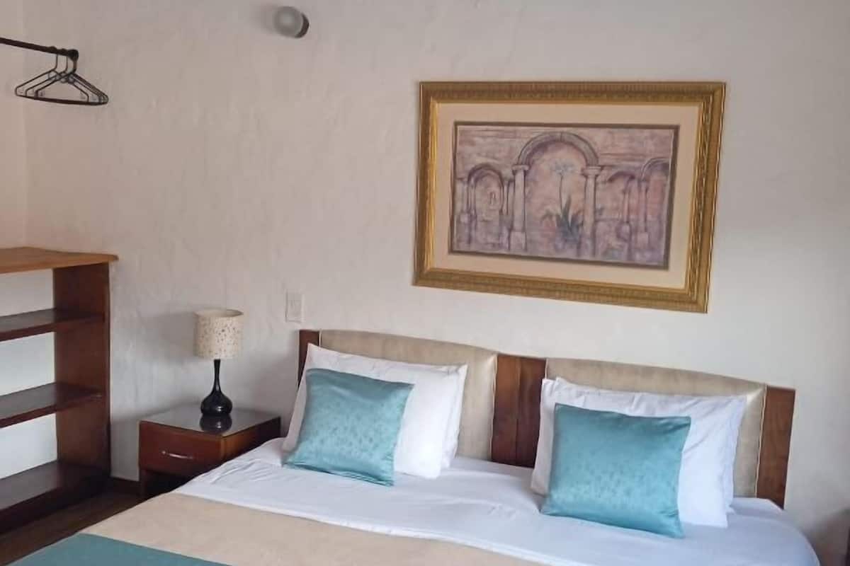 Une chambre à coucher comprenant un lit, une table de chevet, une lampe, une commode en bois et un tableau encadré accroché au mur.