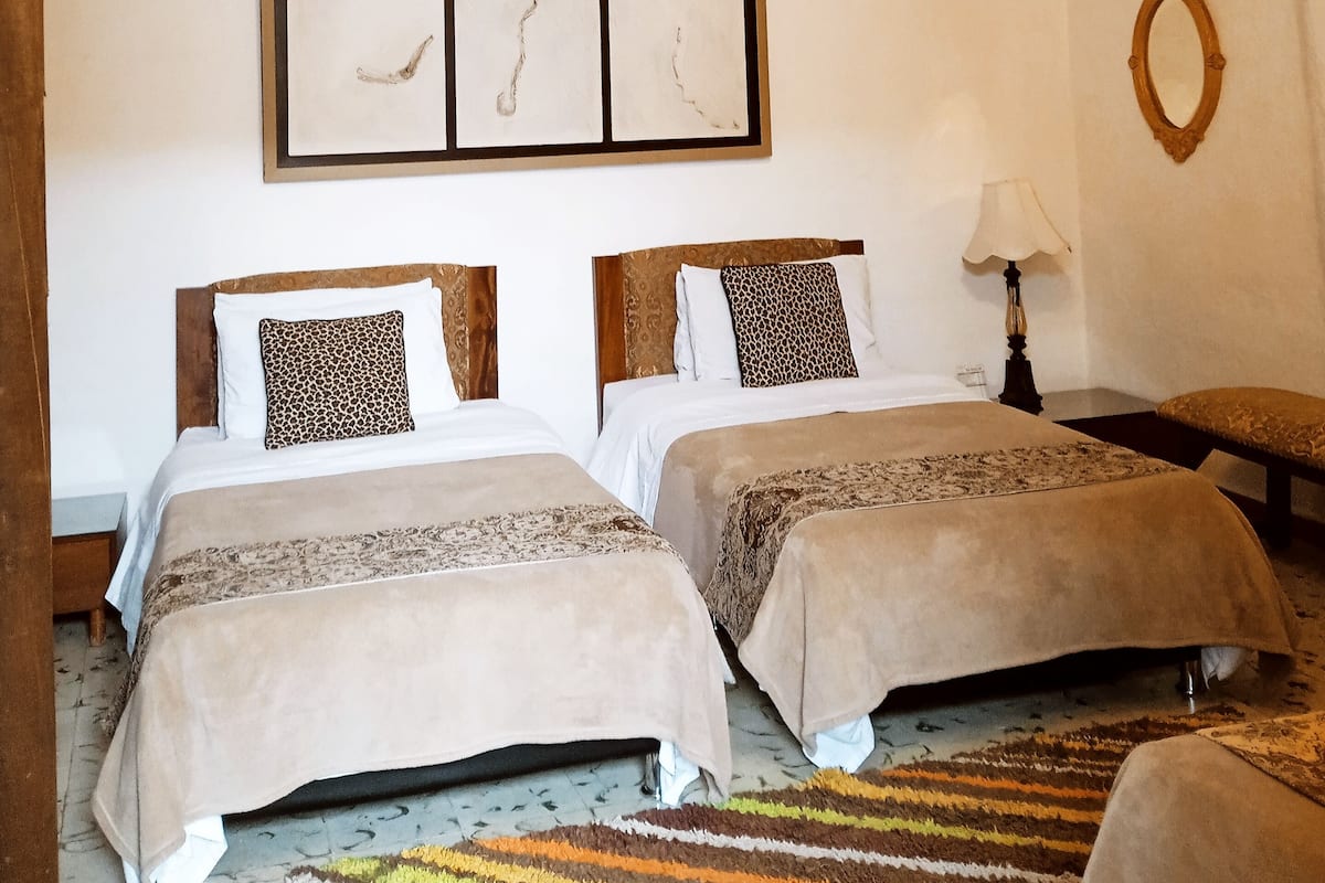 Une chambre avec deux lits, des poutres en bois au plafond, une œuvre d’art encadrée au mur et une petite table avec une lampe.