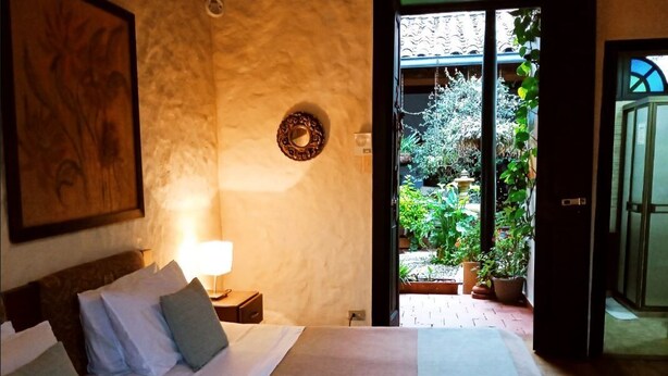 Une chambre à coucher avec un lit, une table de chevet, une lampe fixée au mur et une vue sur un jardin, visible par une porte ouverte.