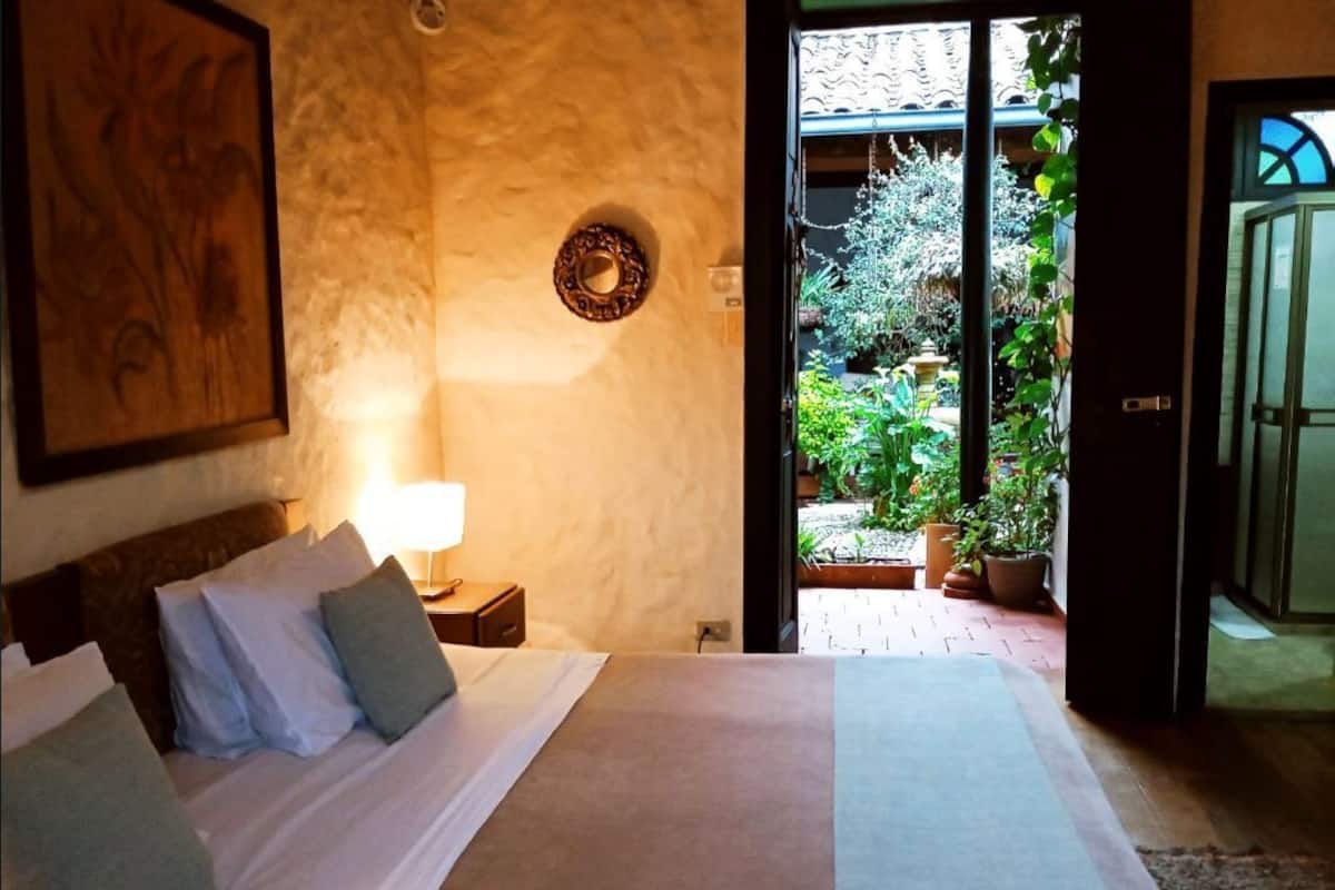 Une chambre à coucher avec un lit, une table de chevet, une lampe fixée au mur et une vue sur un jardin, visible par une porte ouverte.