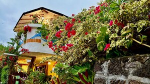 A building with a stone wall and a thatched roof, surrounded by lush greenery and flowering plants. - Casa del Lago Lodging House (Puerto Ayora)