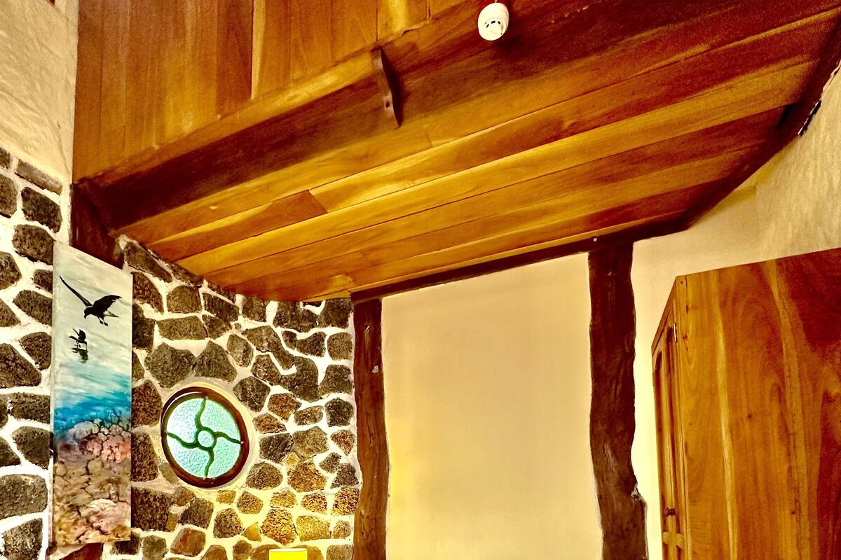 Een slaapkamer met een stenen muur, houten balken in het plafond, een bed met grijze beddengoed en een nachtkastje met een lamp.