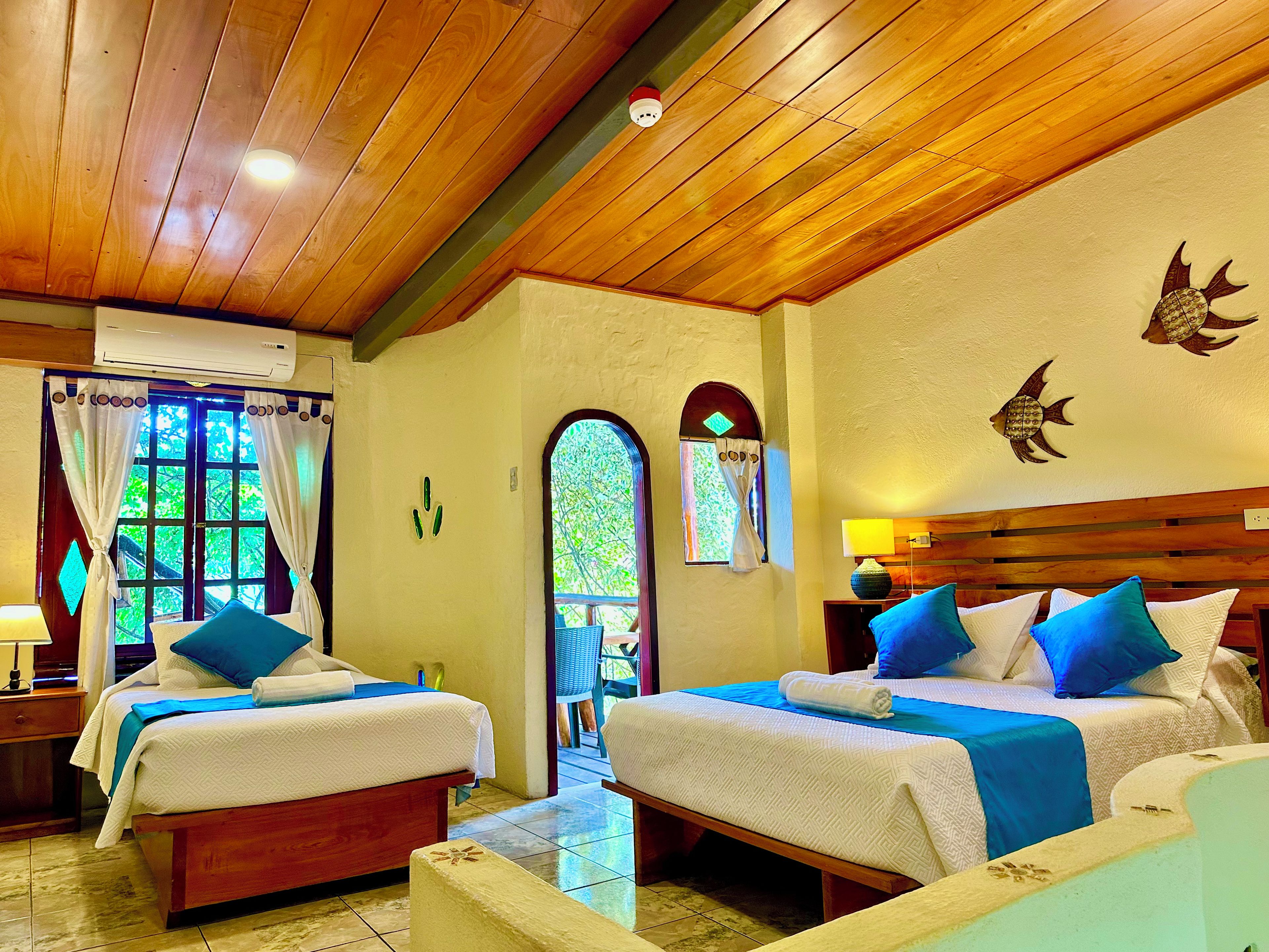 A hotel room with two beds, a wooden ceiling, a window with curtains, a wall-mounted air conditioner, and a small table with a lamp.