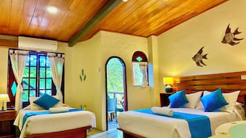 A hotel room with two beds, a wooden ceiling, a window with curtains, a wall-mounted air conditioner, and a small table with a lamp.