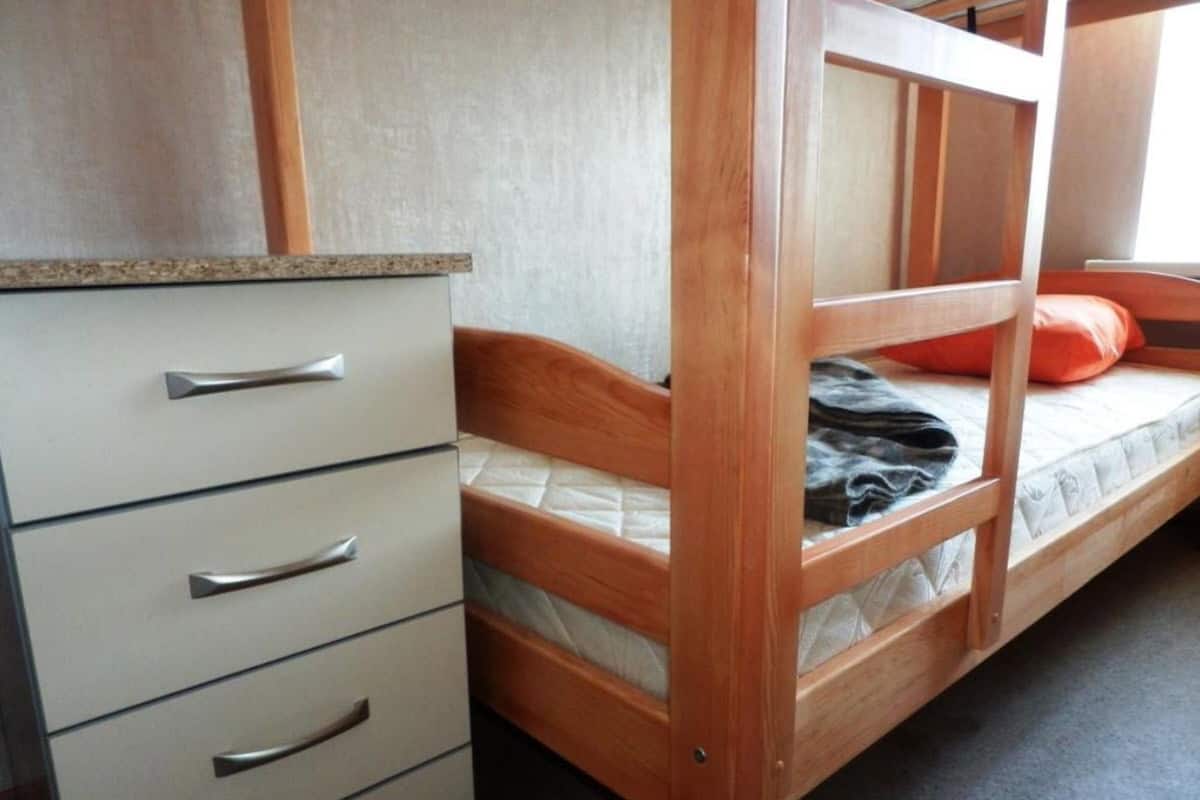 A bunk bed with a wooden frame, a mattress, and an orange pillow. There is a white dresser with silver handles to the left.