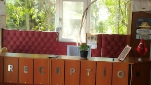 A reception area with a wooden desk labeled "RECEPTION," a red cushioned bench, a potted plant, and a window with a view of greenery. - Gelibolu Kocoglu Hotel (Gallipoli)