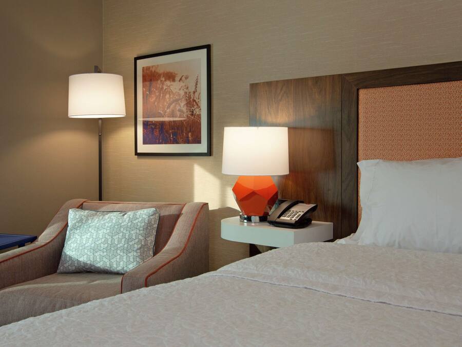 A hotel room with a bed, a chair, a nightstand with a lamp, and a painting on the wall.