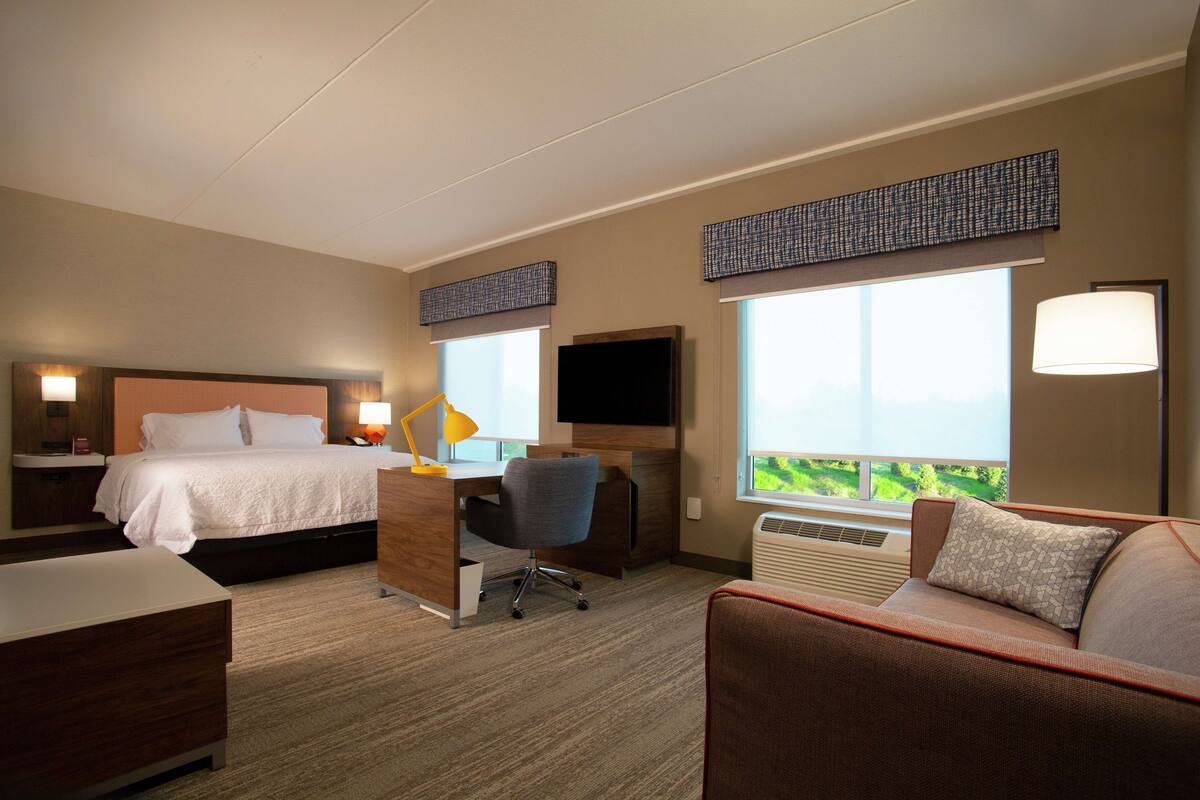 Habitación de hotel con cama, escritorio, silla, sofá, televisor y ventanas amplias.
