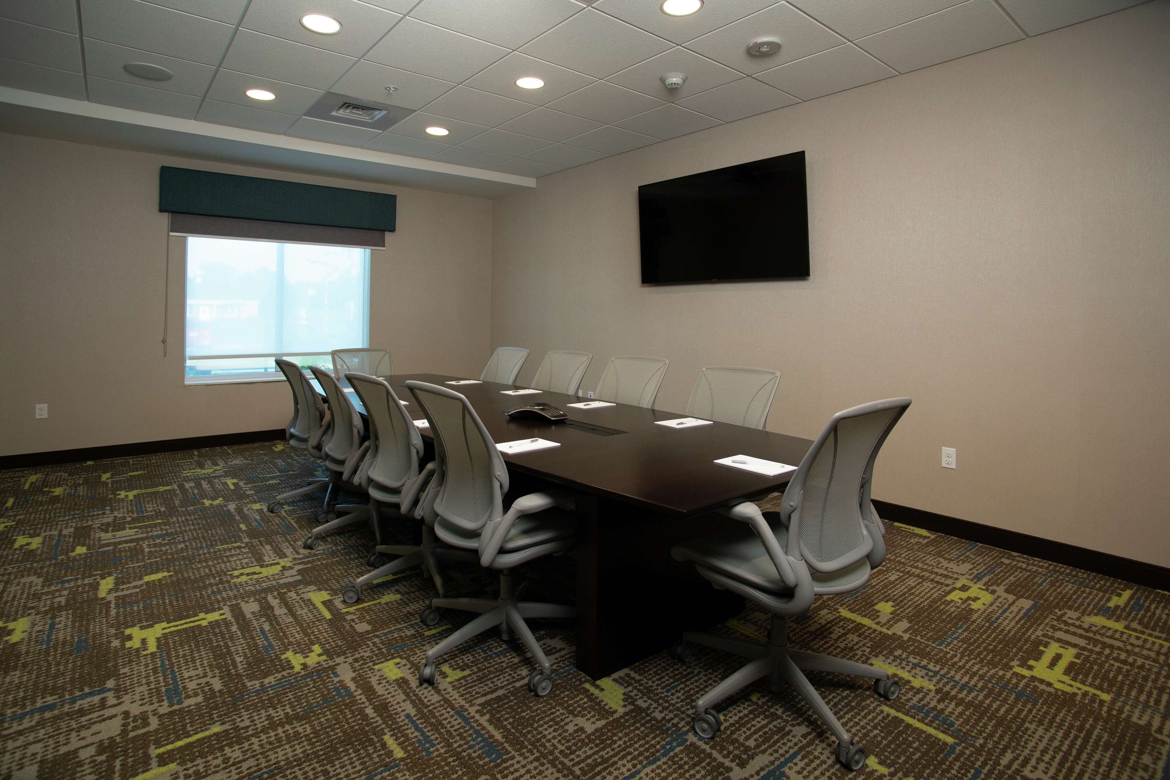 A conference room with a long table, multiple chairs, a large window, and a flat-screen TV on the wall.