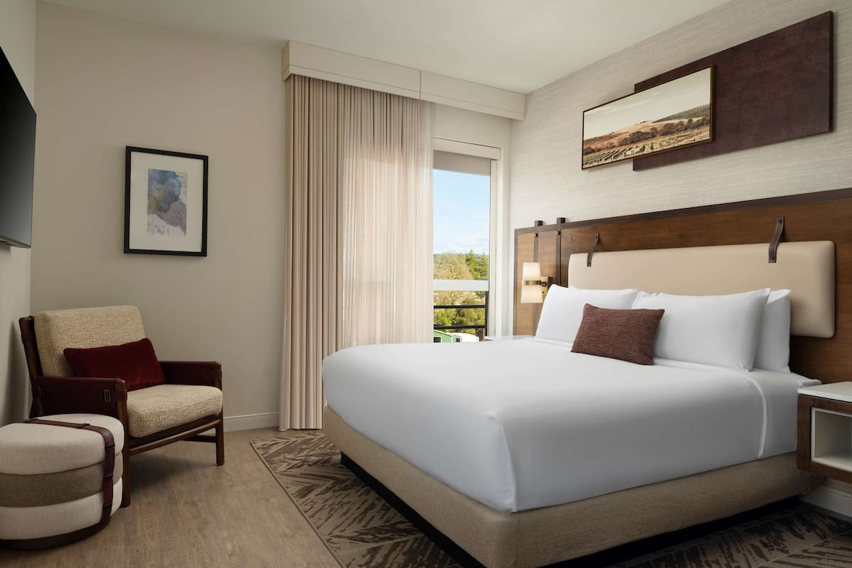 Ein Hotelzimmer mit einem großen Bett, einem Sessel, einem Schreibtisch und einem Fenster mit Vorhängen.