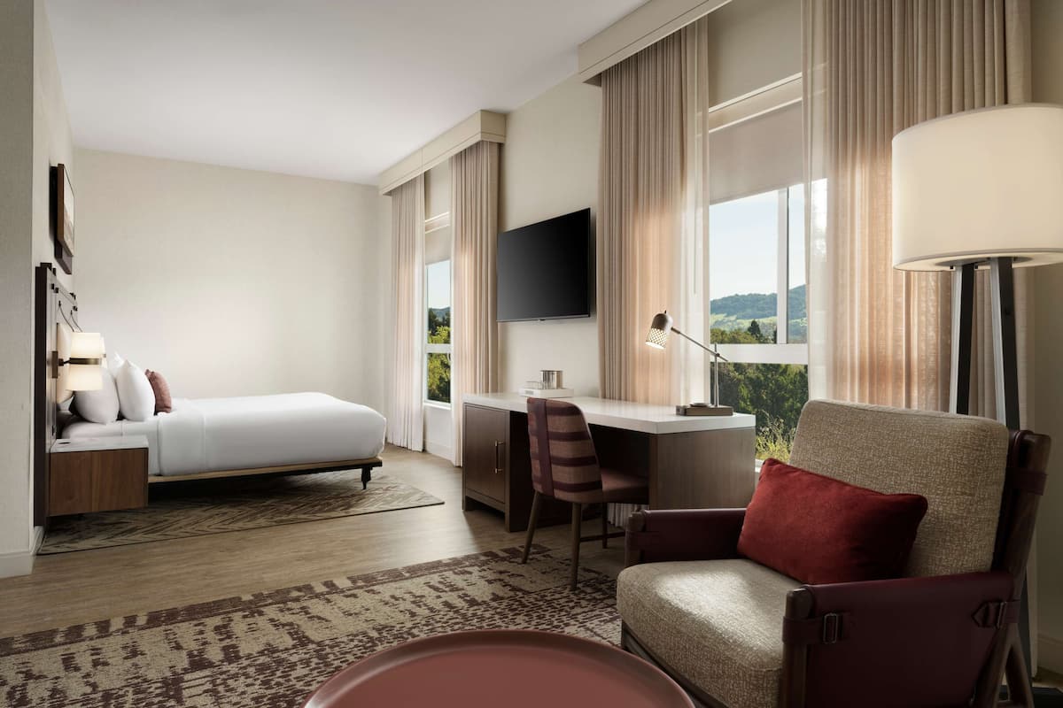 Ein Hotelzimmer mit Bett, Schreibtisch, Stuhl, Fernseher und großen Fenstern mit Vorhängen.
