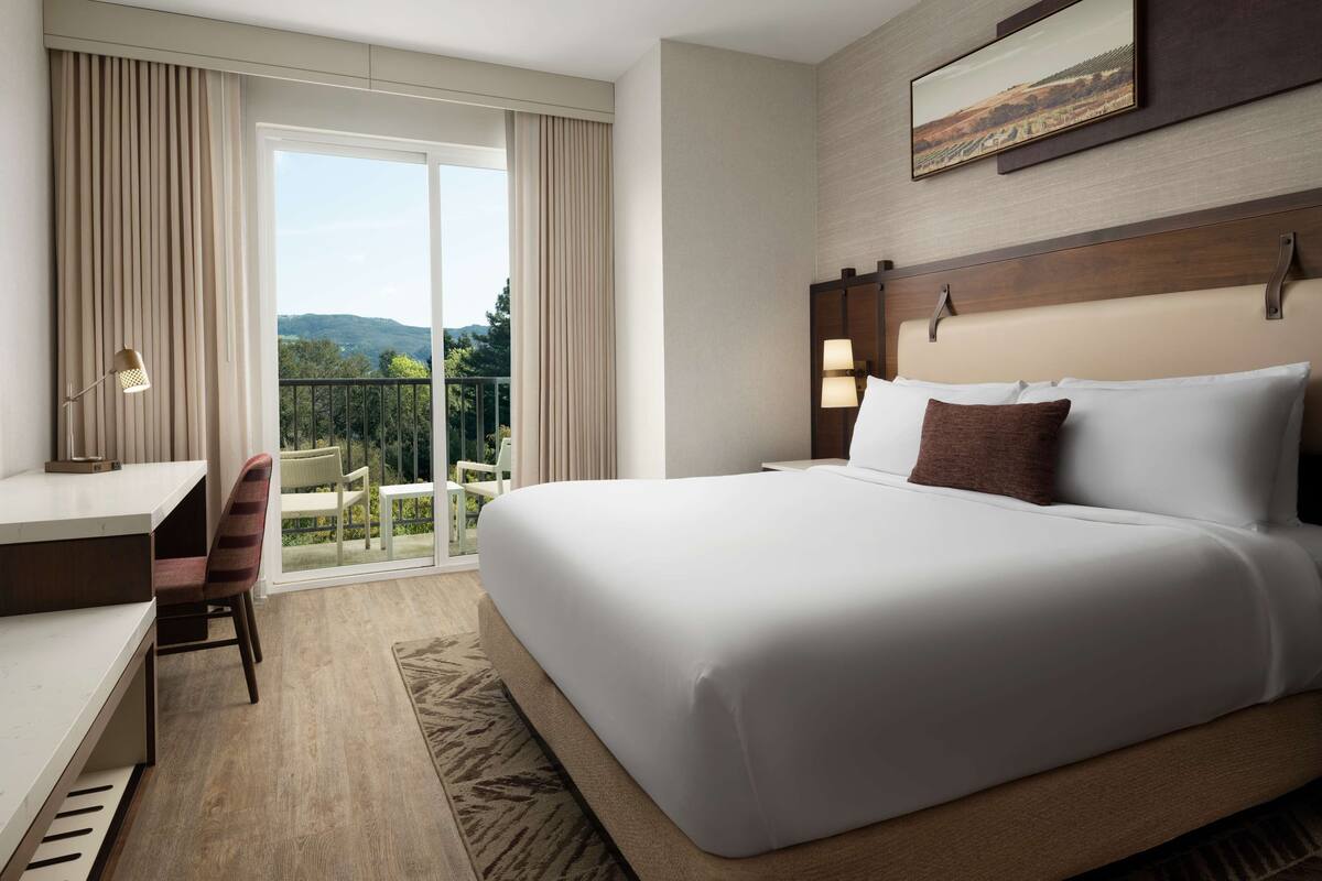 Ein Hotelzimmer mit einem großen Bett, einem Schreibtisch und einem Sessel. Ein Balkon bietet einen Ausblick ins Grüne.