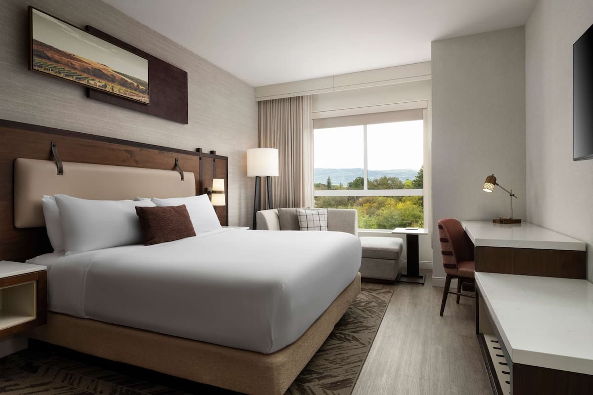 Ein Hotelzimmer mit einem großen Bett, einem Schreibtisch, einem Sessel und einer Lampe, mit Blick ins Freie.