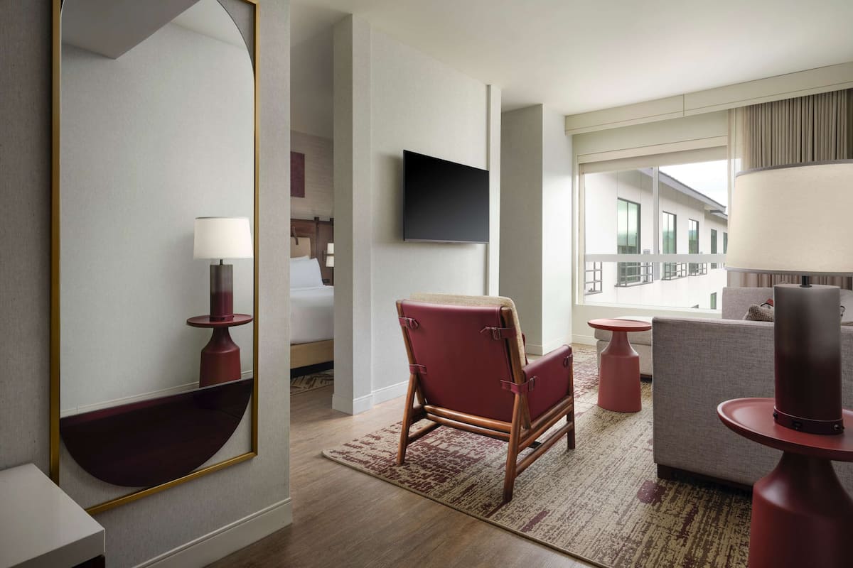 Ein modernes Hotelzimmer mit einem großen Spiegel, einem Flachbildfernseher, einem roten Ledersessel, einem grauen Sofa und einer roten Tischlampe.