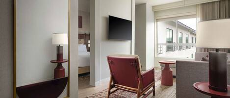 Ein modernes Hotelzimmer mit einem großen Spiegel, einem Flachbildfernseher, einem roten Ledersessel, einem grauen Sofa und einer roten Tischlampe.