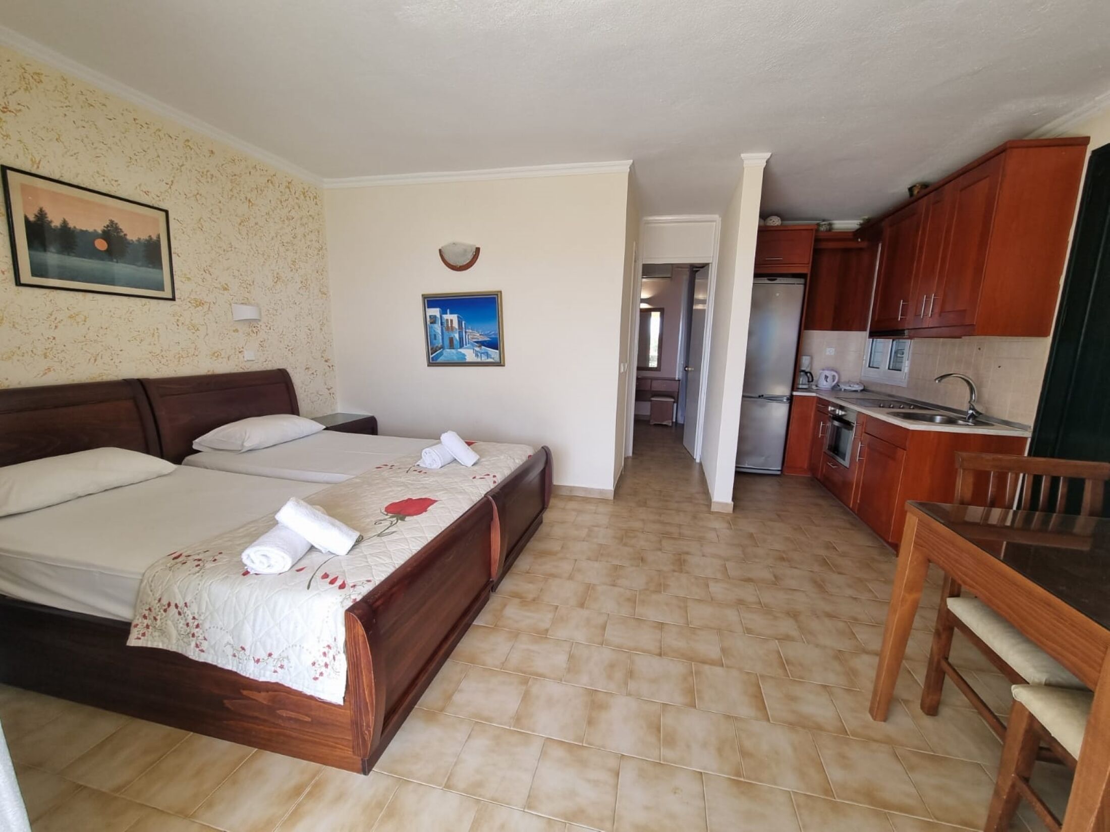 A hotel room with a bed, a small desk, a chair, a kitchenette, and a bathroom visible through an open door.