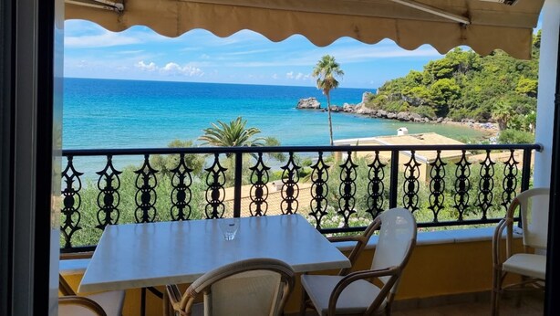 Ein Balkon mit Tisch und Stühlen, der Blick auf einen Strand mit klarem, blauem Wasser und einer felsigen Küste gewährt.